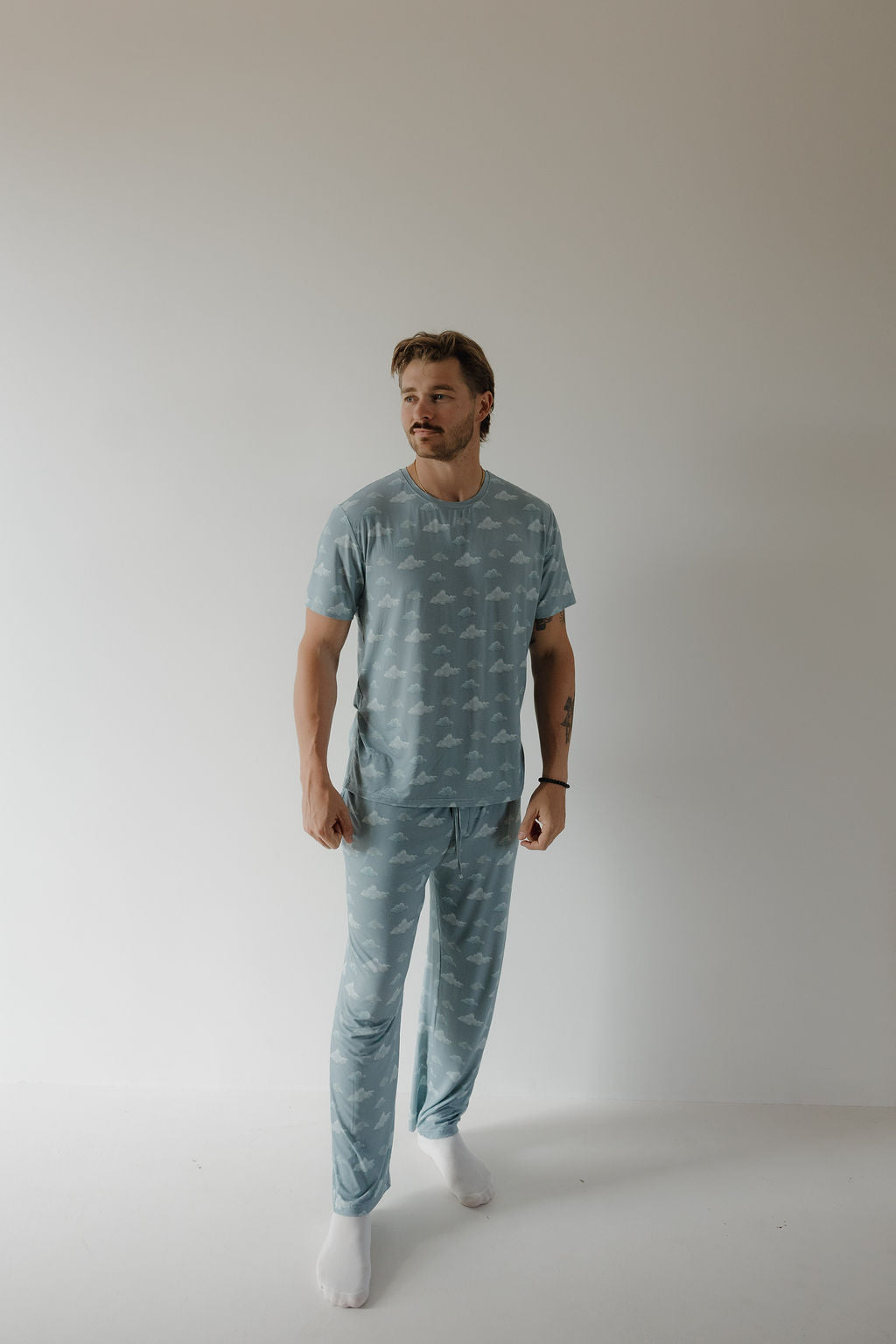 A man with light brown hair, a mustache, and a light beard stands against a plain background, wearing the Kendy 3 Men's Short Sleeve Bamboo Pajama | Duke in light blue with white clouds and white socks.