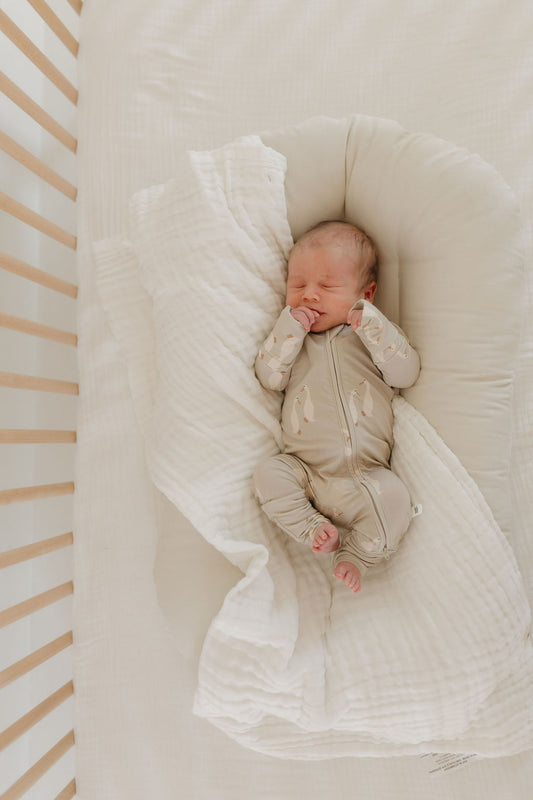 A newborn sleeps peacefully on a cushioned pad inside a crib with wooden slats, wearing Silly Goose Bamboo Zip Pajamas by forever french baby. A crinkled white blanket partially covers the infant.