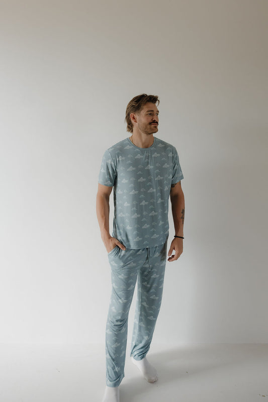 A man with light brown hair and a mustache stands in a minimal, light-filled room wearing the Kendy 3 Men's Short Sleeve Bamboo Pajama | Duke, featuring a breathable white patterned fabric and white socks, hands in pockets, gazing to the side.