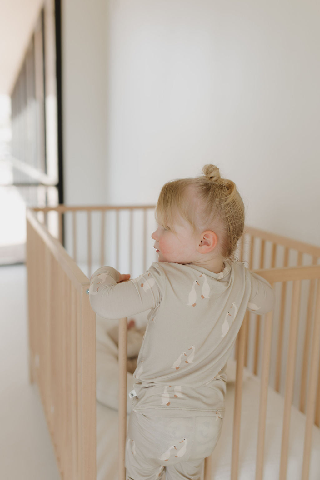 A blonde toddler in a small bun stands in a light wooden crib, facing away. Wearing "Bamboo Two Piece Pajamas" by Forever French Baby, the child blends seamlessly into the minimalist room with white walls and a large window.