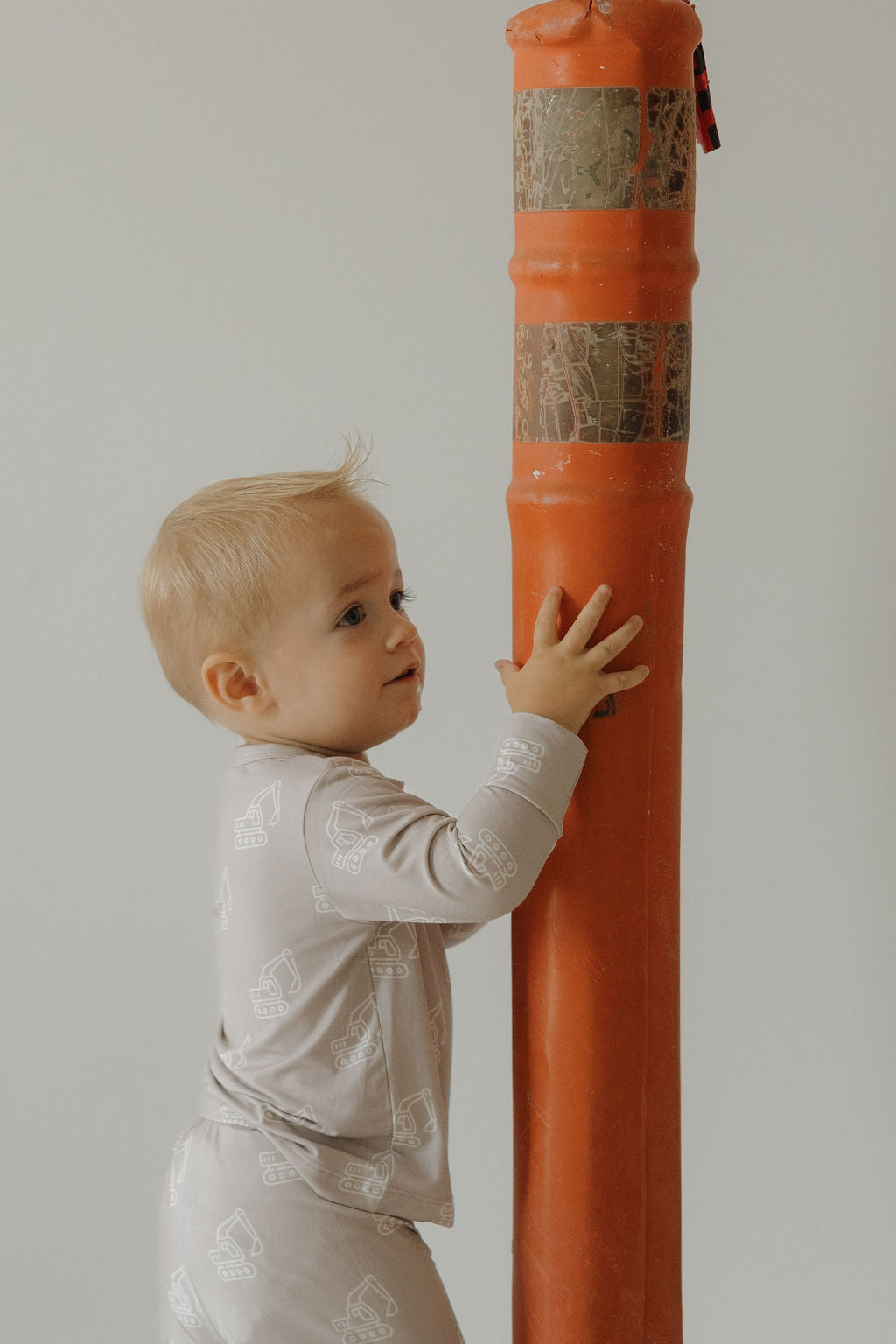 A young child wears the forever french baby Child Sleepy Time Set | Excavator, standing by an orange pillar and gazing upward with curiosity. The neutral background emphasizes the set’s hypo-allergenic comfort and light bamboo fabric.