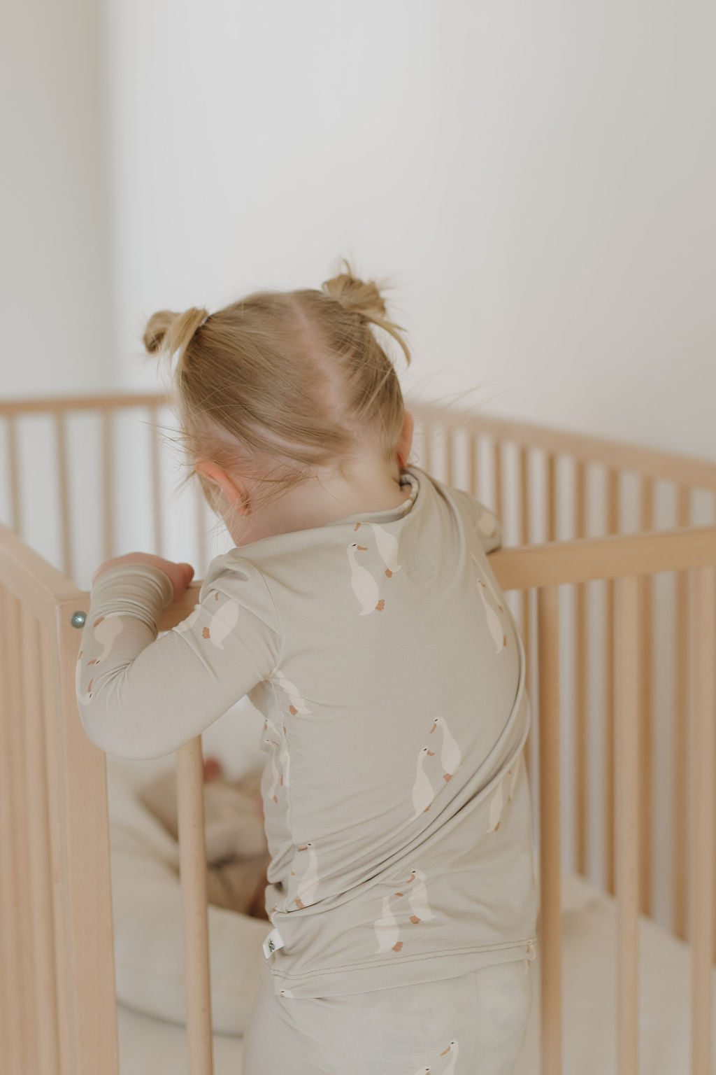 A young child with two small ponytails stands in a light wooden crib, peering down. Dressed in Bamboo Two Piece Pajamas by Silly Goose from forever french baby, they exude comfort and style. The softly blurred background adds to the scene's calm and neutral tone.