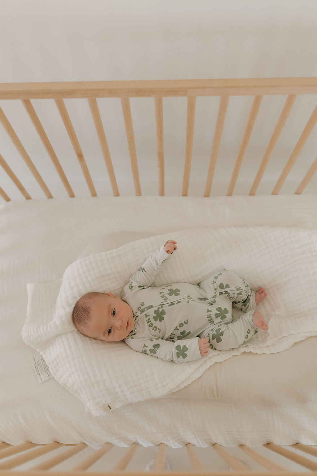 A baby lies in a wooden crib with slatted sides on a light-colored blanket, wearing the charming Bamboo Zip Pajamas | Lucky You by forever french baby. The little one gazes upwards, snug in breathable fabric ideal for sensitive skin.