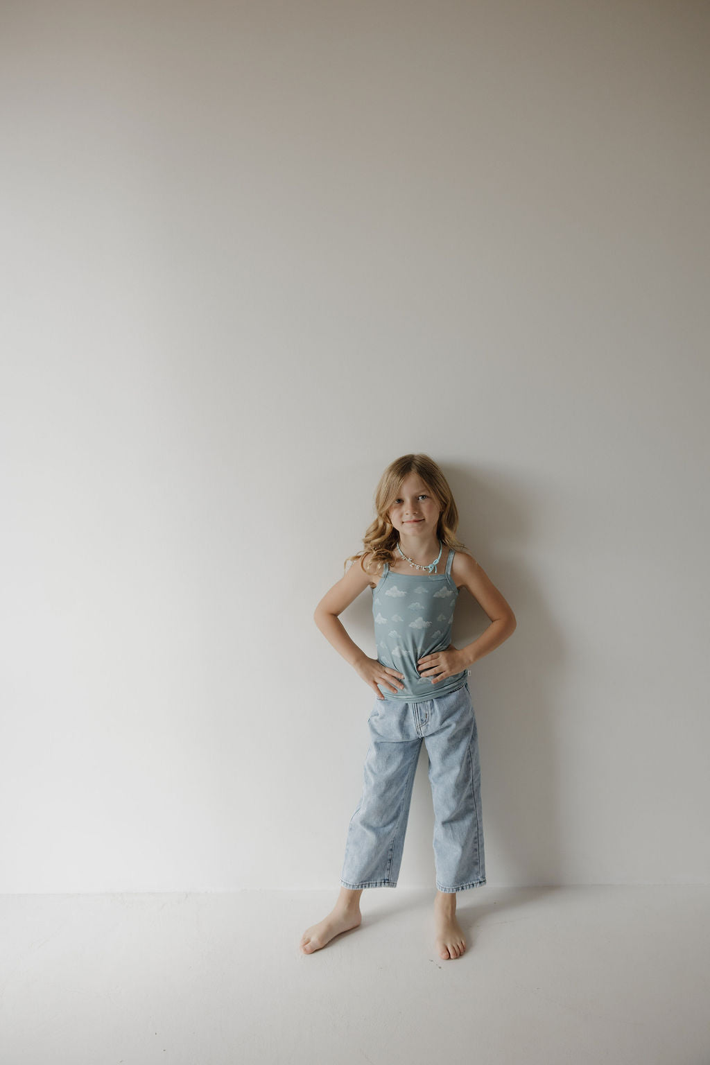 A young girl with long blonde hair stands barefoot against a plain wall, wearing the Kendy 3 Toddler Bamboo Cami Set | Duke—a light blue breathable tank top and loose-fitting jeans—with her hands on her hips and a slight smile.