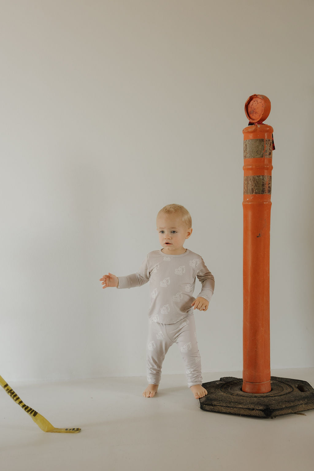 A toddler in the forever french baby Child Sleepy Time Set | Excavator stands indoors beside a tall orange traffic post on a white floor, with part of a yellow and black striped object visible on the left.