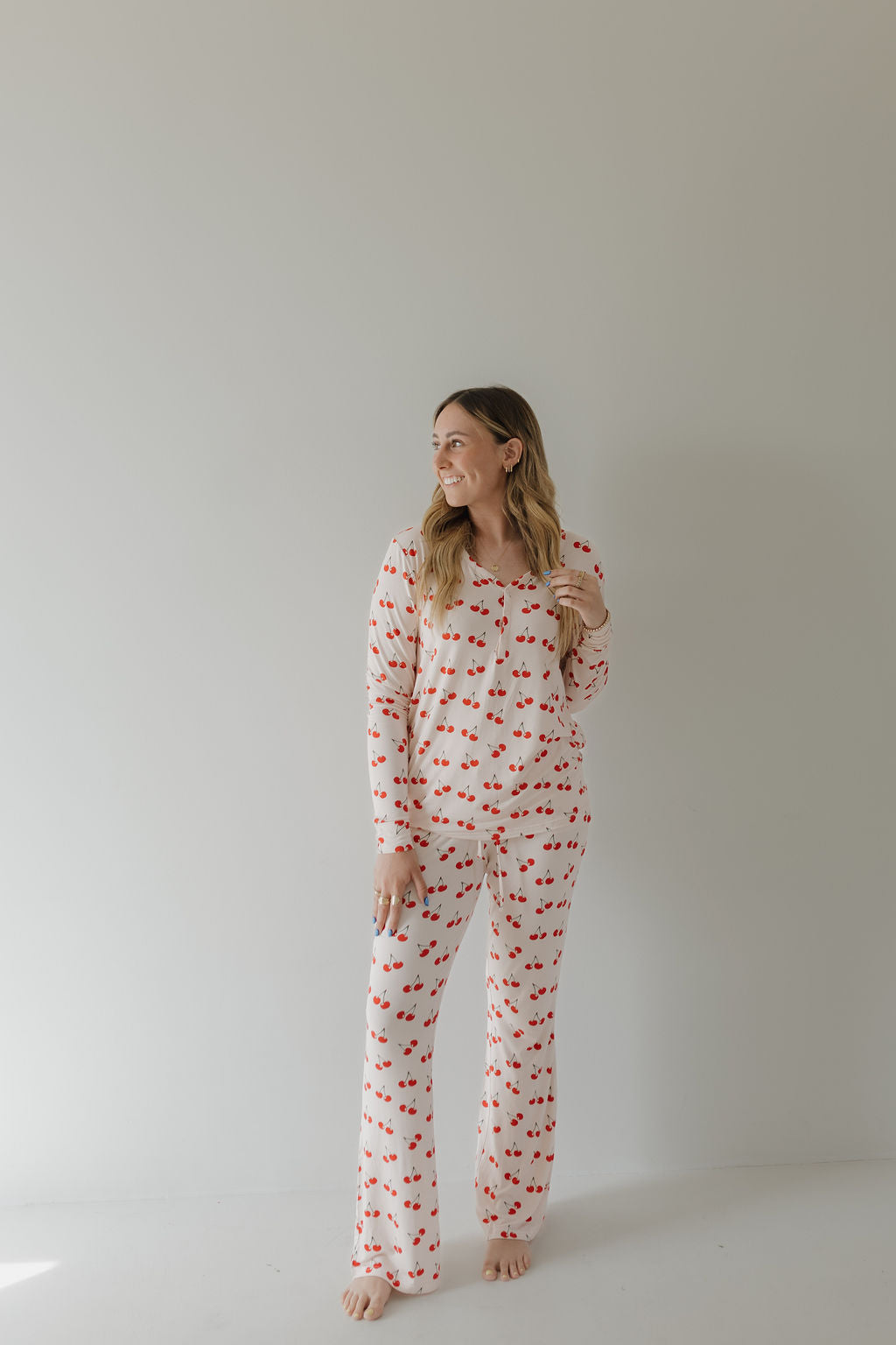 A person with long hair stands barefoot against a plain background, wearing forever french baby's Women's Bamboo Pajama in Cherry Orchard design. With a relaxed posture and gentle smile, they look to the side, exuding an air of year-round comfort.