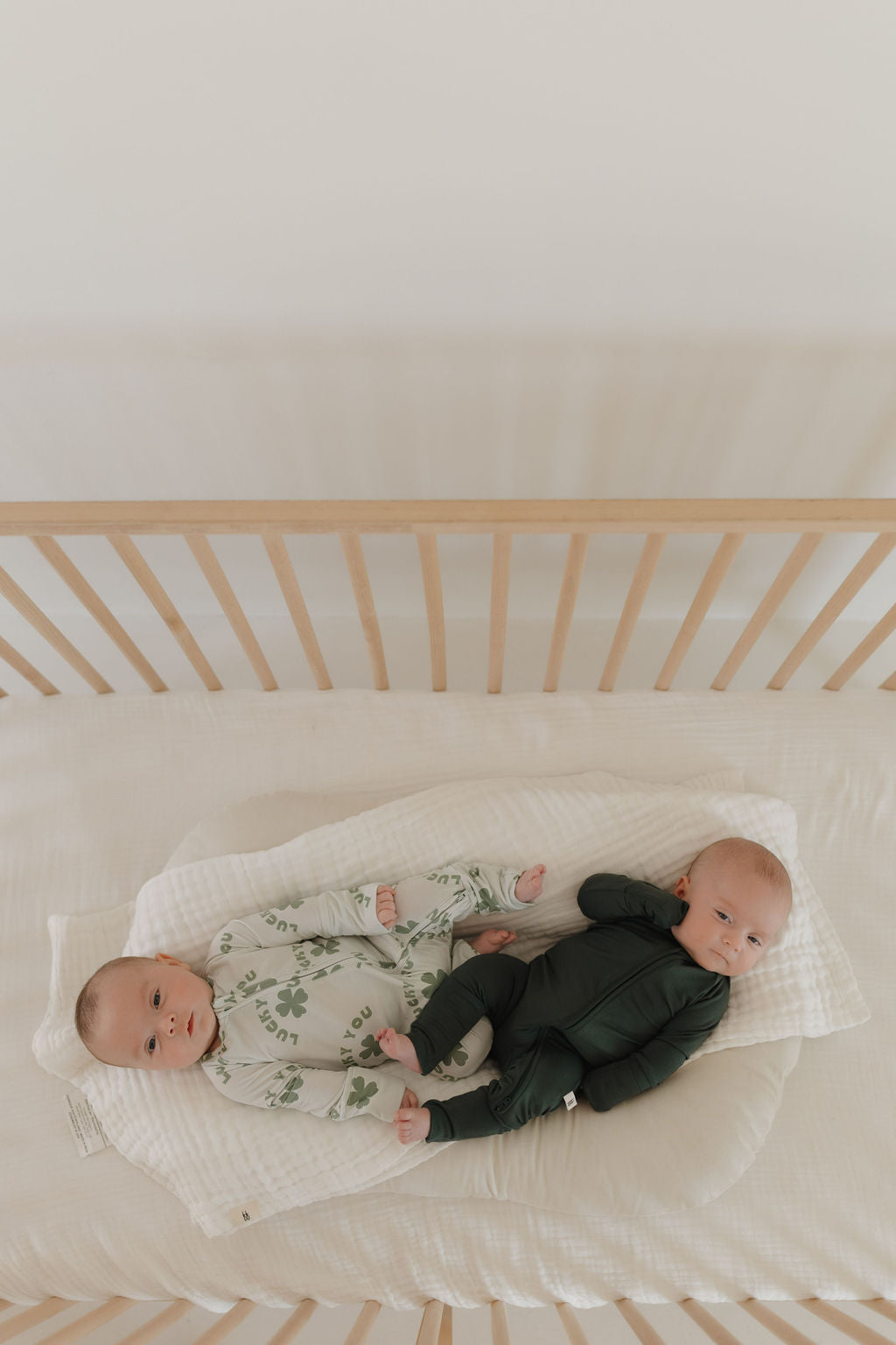 Two babies rest in a light wood crib. One is in a green outfit with nature print, and the other wears forever french baby Bamboo Zip Pajamas in Emerald, praised for being breathable and hypoallergenic. They lie on a padded white blanket, gazing upwards.