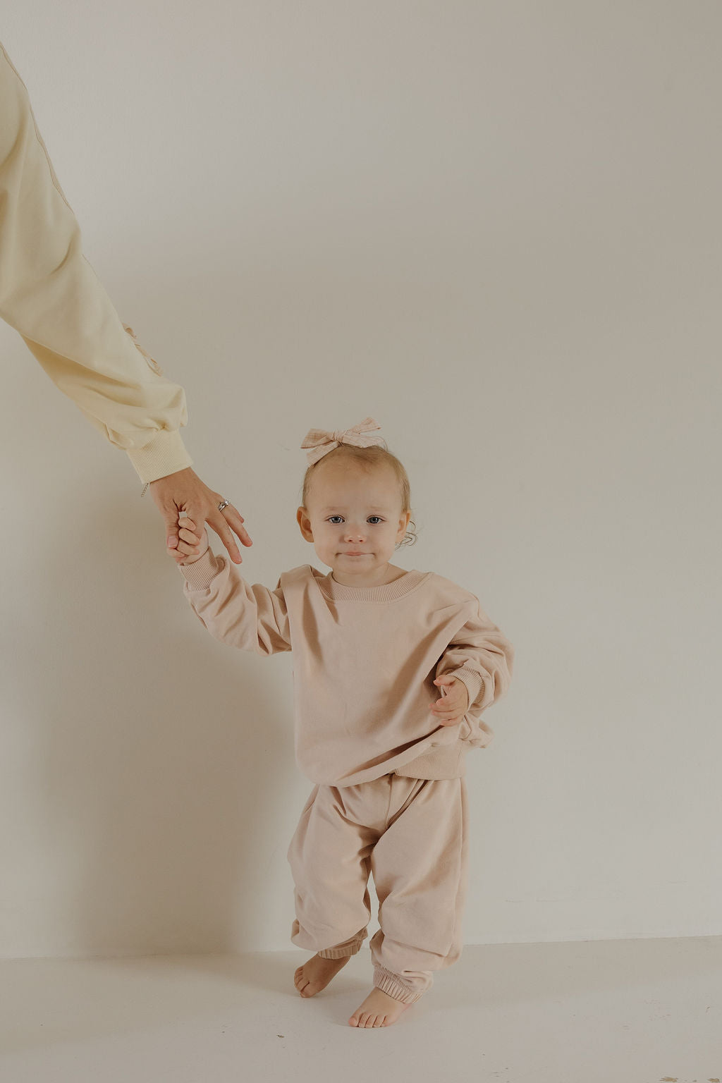 A toddler in the forever french baby Child Sweat Set in Cashew with a matching bow holds an adult’s hand, ready for everyday adventures against a plain, light background.