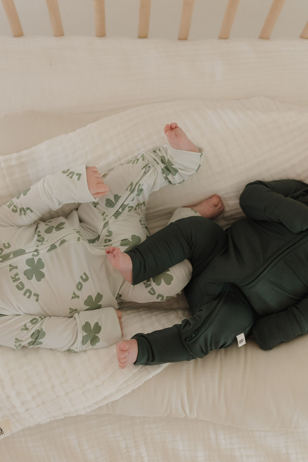 Two babies snuggle in a crib, wearing "forever french baby" Bamboo Zip Pajamas. One outfit is adorned with green shamrocks and "lucky" on white fabric, while the other is solid emerald. These breathable pajamas ensure comfort on the plush white mattress—a cozy haven for restful sleep.