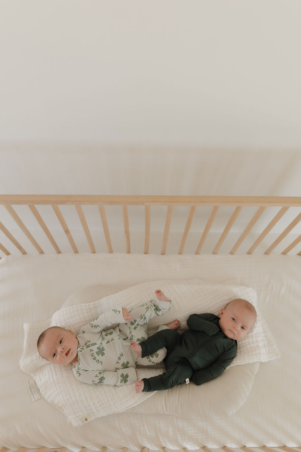 Two babies lie in a crib with wooden rails, resting on a white blanket. One wears light-patterned pajamas, and the other is dressed in Emerald Bamboo Zip Pajamas by forever french baby. The cozy setting features neutral tones as both look up peacefully on a soft mattress.