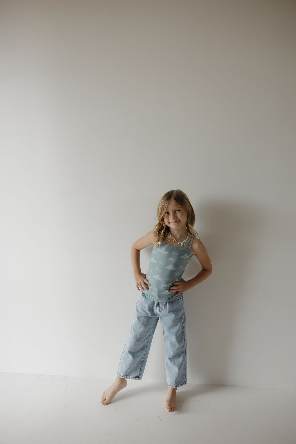 A young girl with long, wavy blonde hair stands barefoot against a white wall, smiling slightly with her hands on her hips in the Kendy 3 Toddler Bamboo Cami Set | Duke—a breathable, light blue tank top and loose-fitting jeans.