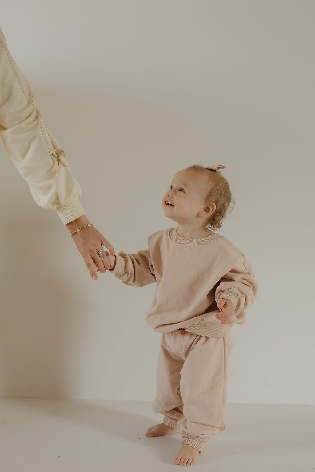 A small child smiles up while wearing the forever french baby Child Sweat Set | Cashew, holding an adult’s hand against a plain, light background—perfect for everyday adventures.