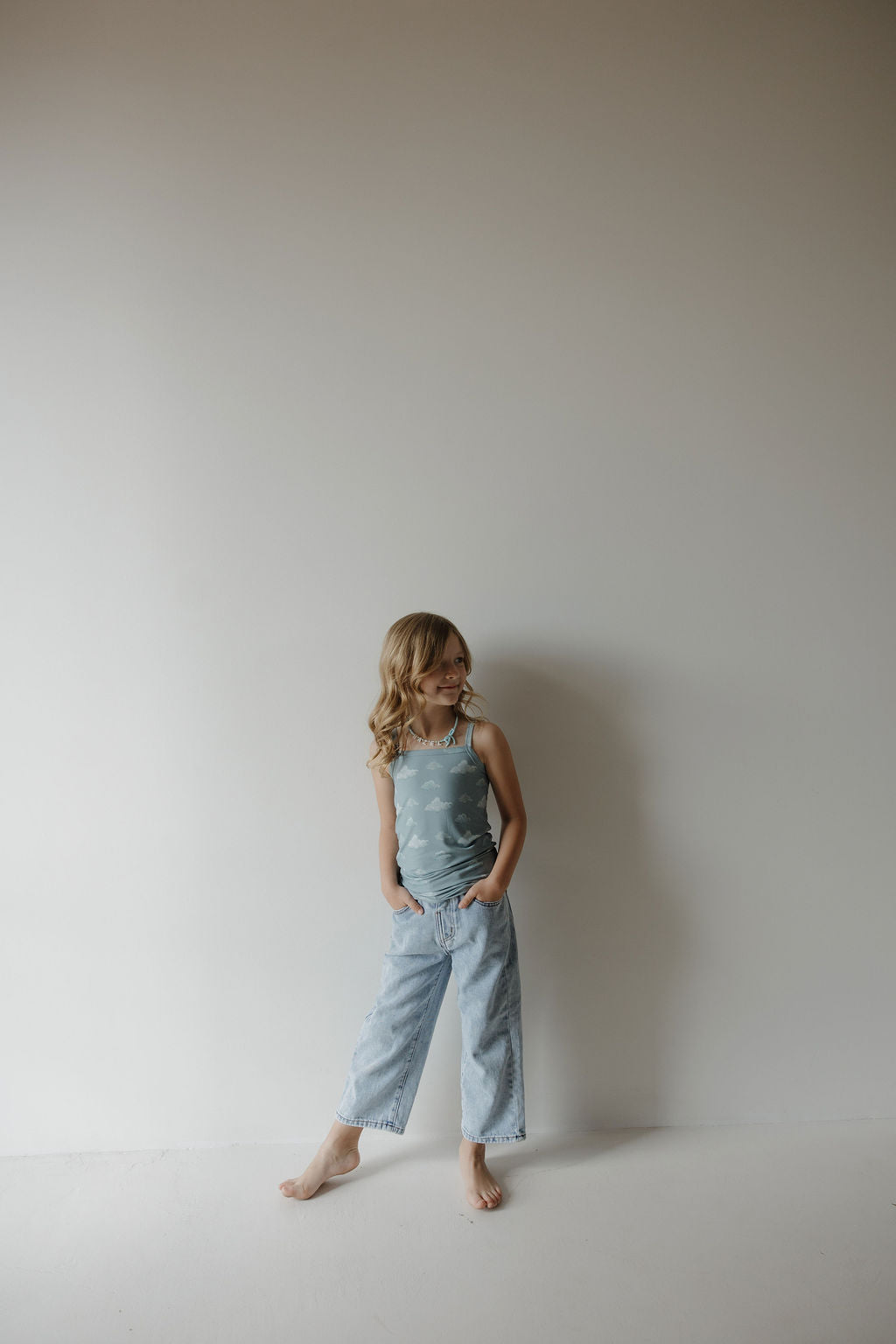 A young girl with long blonde hair stands barefoot against a plain wall, wearing the Kendy 3 Toddler Bamboo Cami Set in Duke, with her hands in her pockets, looking off to the side and smiling.