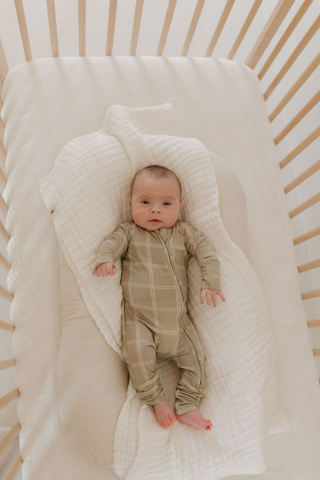 A baby in a wooden crib, swaddled in a white blanket, wears the forever french baby Bamboo Zip Pajamas in Linen Grid. The crib and bedding feature a soft, neutral color palette as the baby looks up with a calm expression.