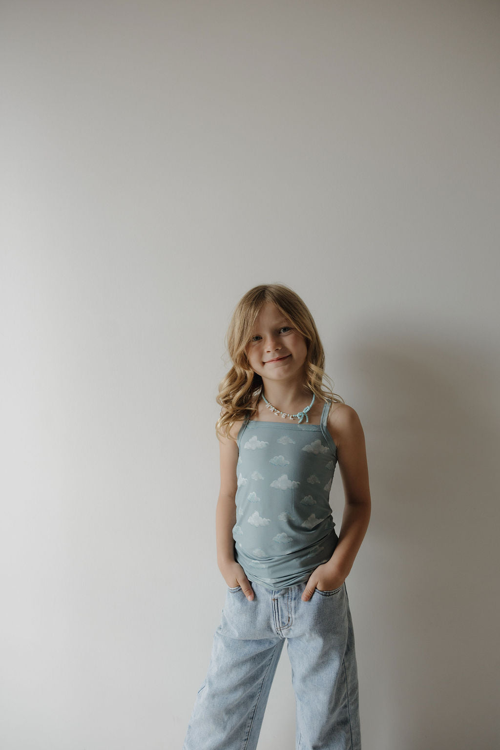 A young girl with long blonde hair, wearing the Kendy 3 Toddler Bamboo Cami Set | Duke, smiles against a light-colored wall. She pairs the soft cami and shorts set with jeans and a colorful beaded necklace, her hands in her pockets.