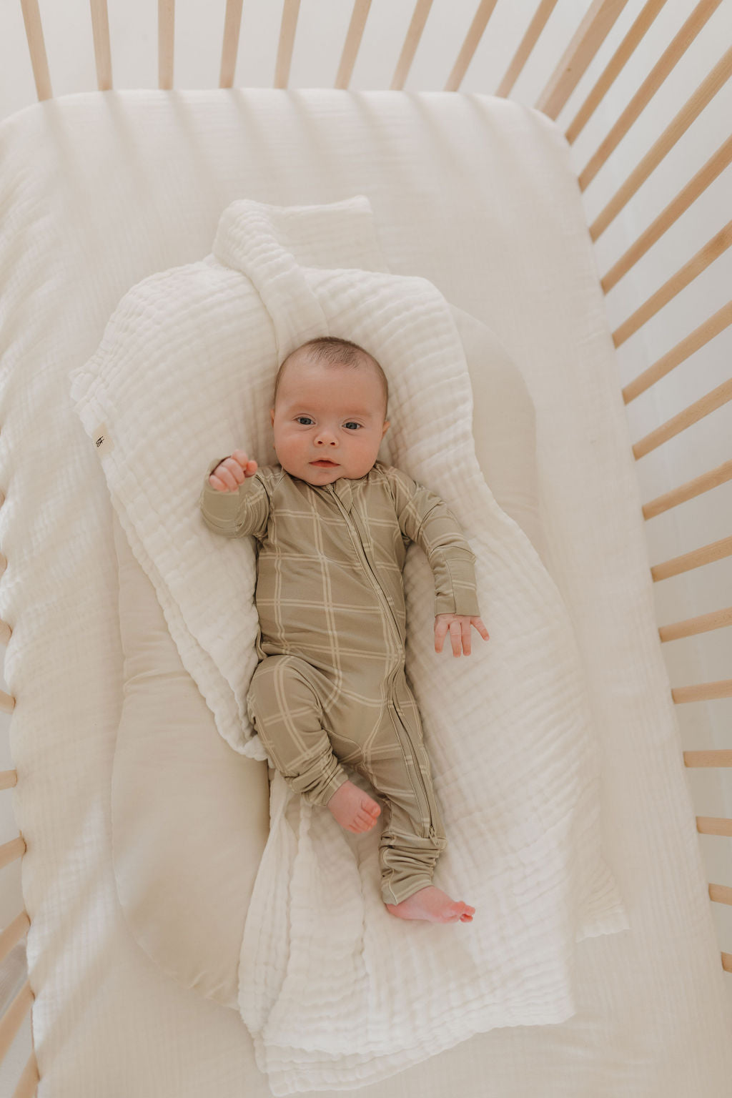 A baby in forever french baby's Bamboo Zip Pajamas | Linen Grid lies on a white quilted blanket inside a softly lit, warm wooden crib, one arm raised and the other at their side.