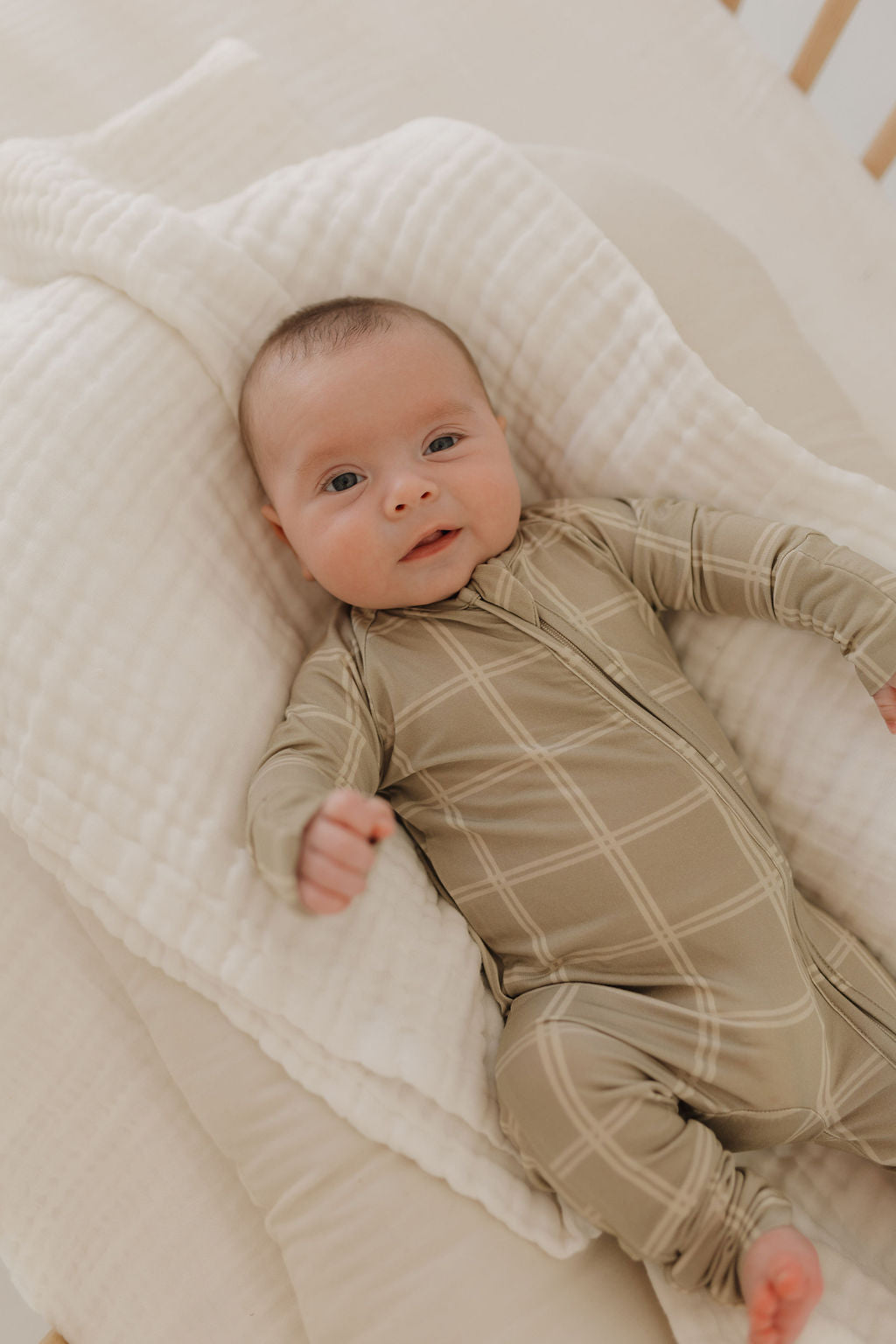 A baby rests on a soft, cream-colored blanket, wearing forever french baby's Bamboo Zip Pajamas in Linen Grid. The baby gazes upward contentedly, surrounded by soothing, neutral tones.