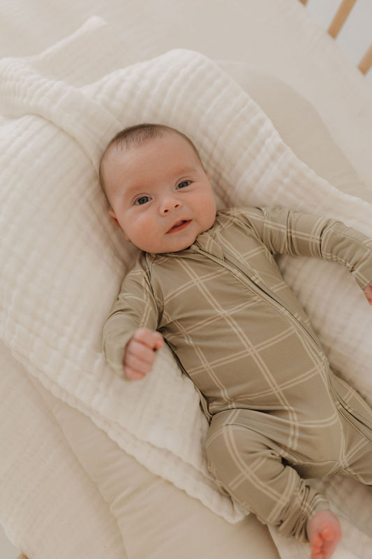 A baby rests on a soft, cream-colored blanket, wearing forever french baby's Bamboo Zip Pajamas in Linen Grid. The baby gazes upward contentedly, surrounded by soothing, neutral tones.