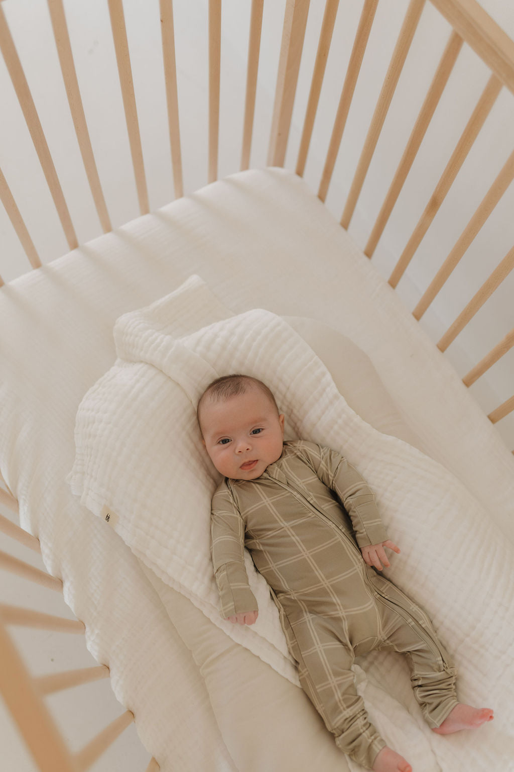 A baby in forever french baby's Bamboo Zip Pajamas | Linen Grid lies on a fluffy white blanket in a wooden crib with light-colored slats, gazing upward calmly.