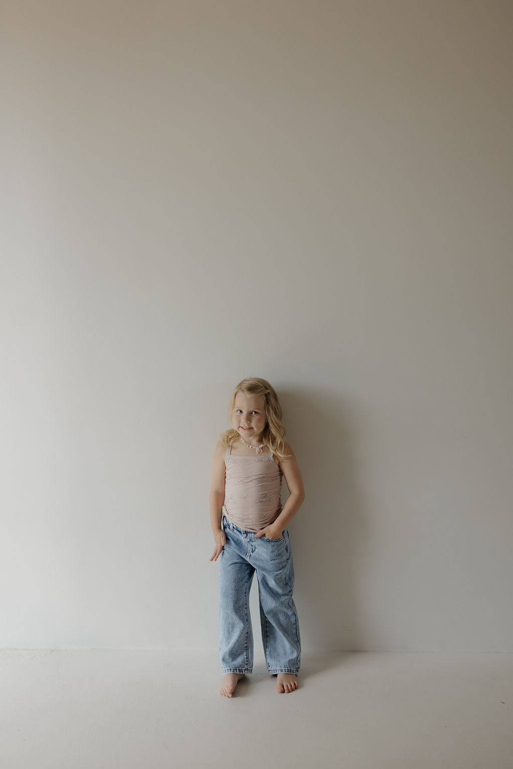 A young girl with blonde hair stands barefoot against a plain light wall, wearing the Kendy 3 Toddler Bamboo Cami Set in Ruby and loose blue jeans, with her hands in her pockets and looking slightly to the side.