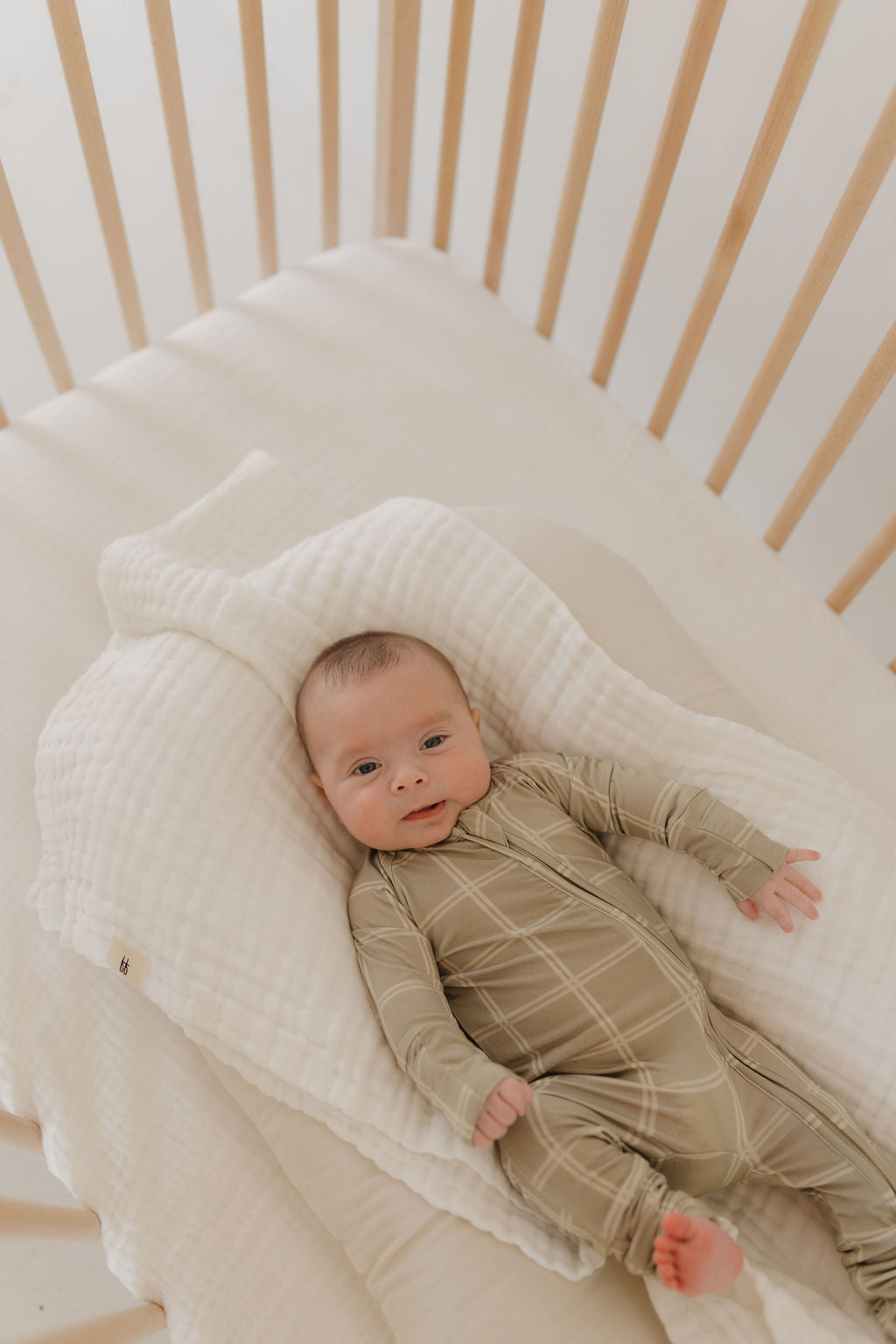 A baby wearing forever french baby's Bamboo Zip Pajamas in Linen Grid lies on a soft, white blanket inside a wooden crib with vertical slats. The baby looks up with a slight smile, creating a calm and cozy atmosphere.