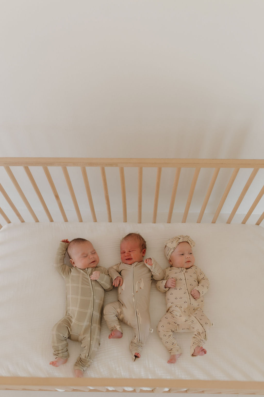 Three babies lie side by side in a wooden crib, each wearing a different onesie: one sports the Bamboo Zip Pajamas in Linen Grid by forever french baby. Another is in solid beige, and the last wears beige with small prints featuring a matching headband. They're set against a plain light-colored wall.