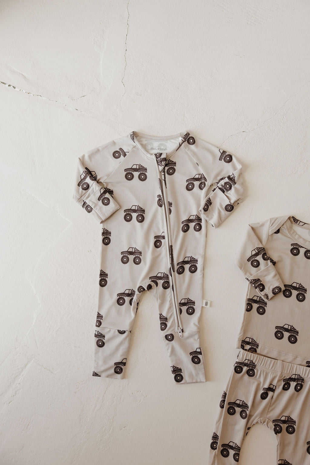 The Bamboo Zip Pajamas | Monster Truck by forever french baby feature a beige long-sleeve romper with a zipper front and black truck print, crafted from breathable fabric. Shown laid flat with matching pants and top partially visible on the right.