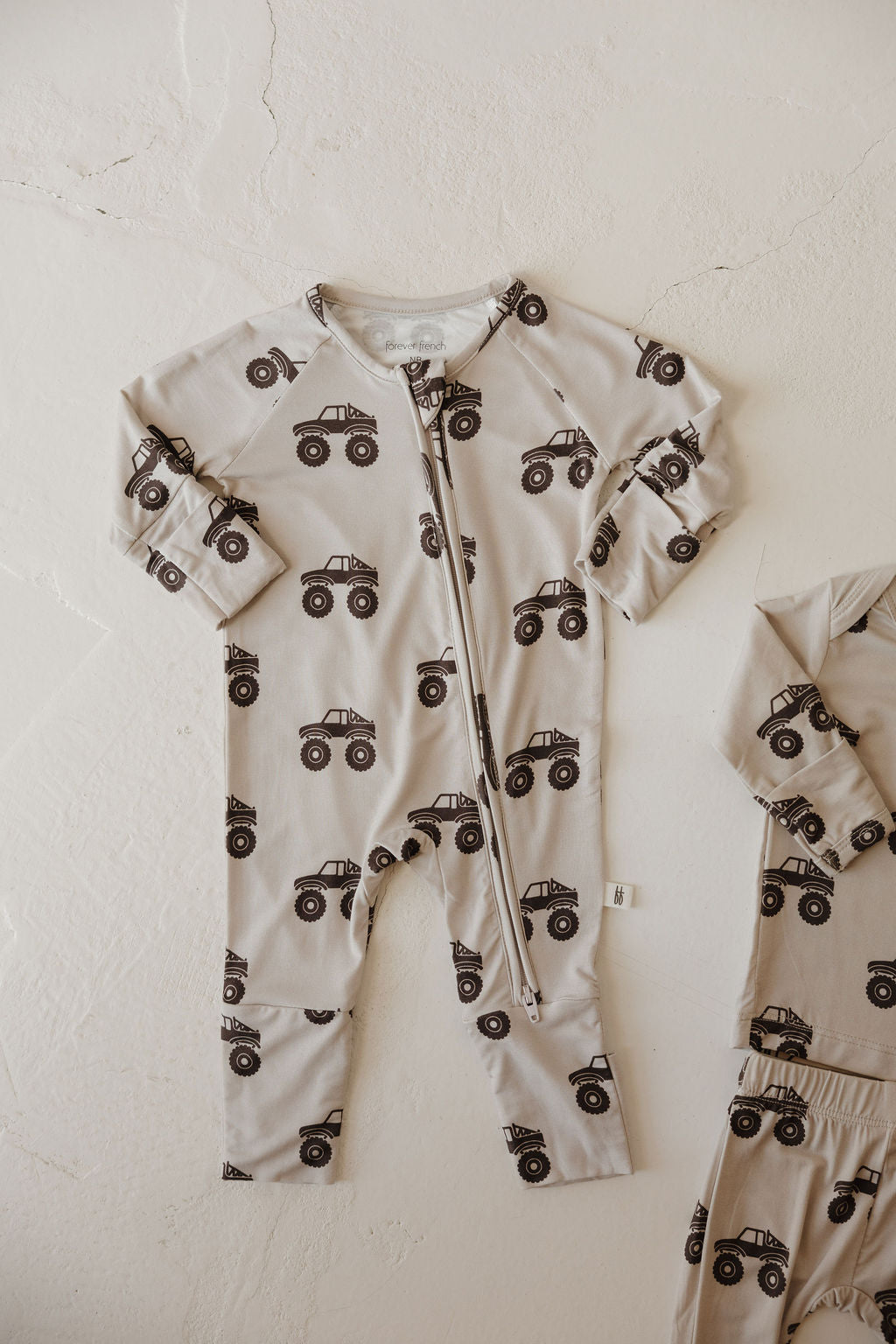 The Bamboo Zip Pajamas | Monster Truck by forever french baby features a beige, breathable fabric with a black monster truck print and zipper front, laid flat on a white surface. A matching sleeve and pant leg are partially visible on the right.