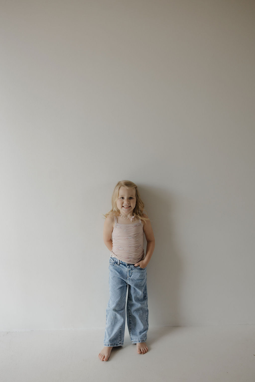 A young child with blond hair stands barefoot by a plain wall, wearing the Kendy 3 Toddler Bamboo Cami Set | Ruby—featuring a sleeveless beige bamboo top and loose blue jeans—with hands in pockets and a neutral look.