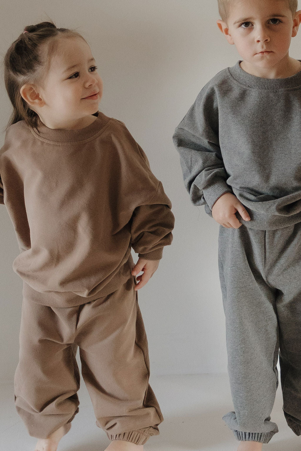 Two young children stand side by side against a plain background, each wearing the forever french baby Child Sweat Set in Charcoal—an ideal choice for creating coordinated family looks.