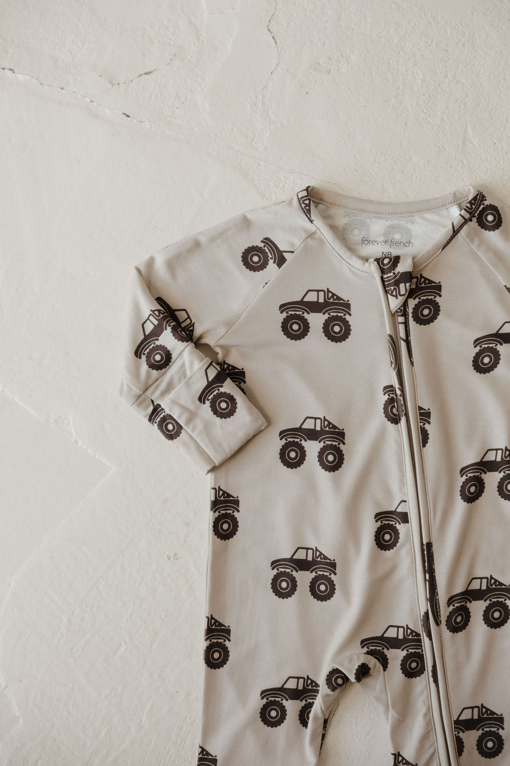 The forever french baby Bamboo Zip Pajamas | Monster Truck is a beige baby onesie with a zipper front, made from breathable fabric and decorated with dark brown monster trucks, shown flat on a light textured surface.