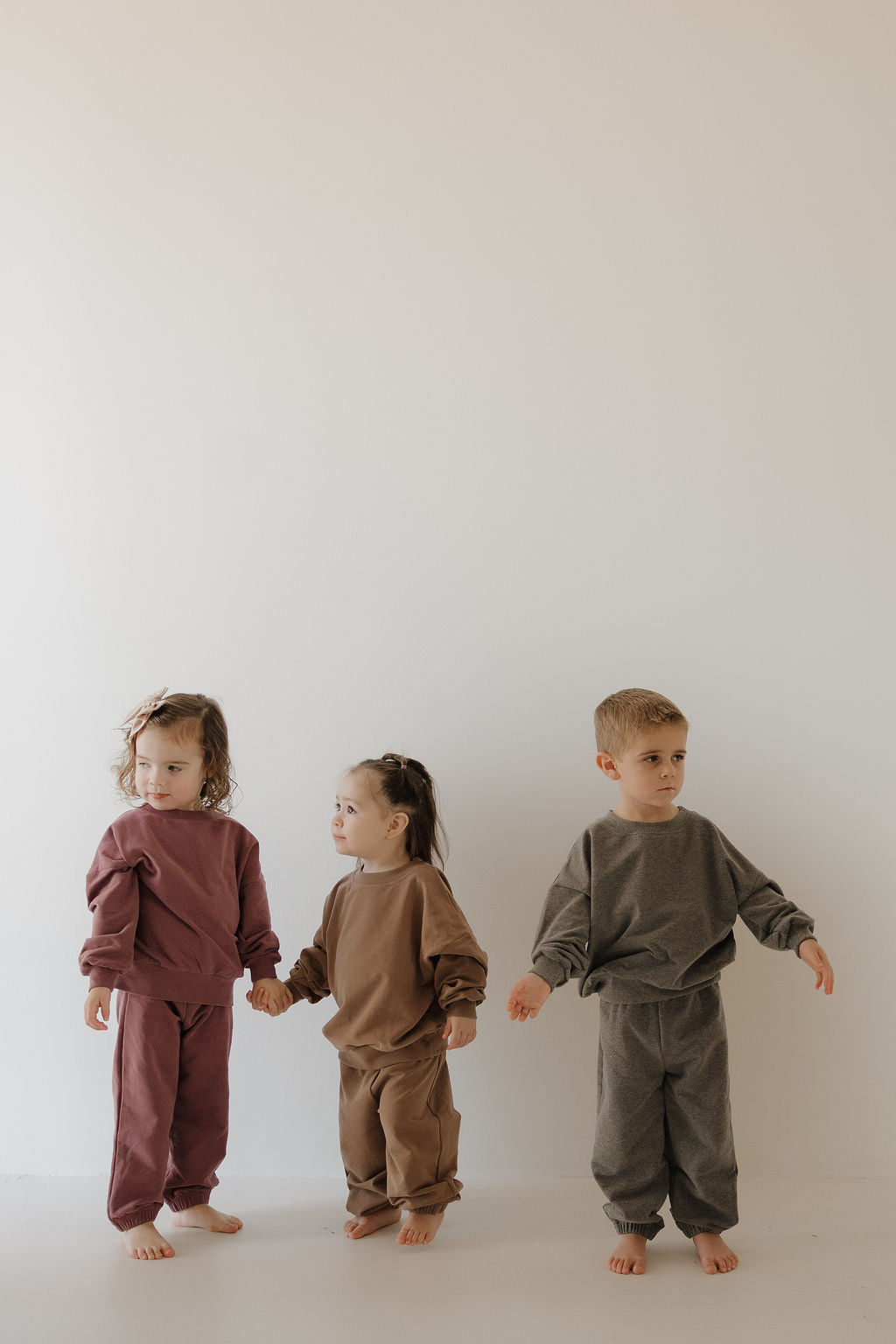Three young children stand barefoot against a light wall, each wearing the forever french baby Child Sweat Set | Twilight. The coordinated sets feature solid colors in maroon, brown, and gray as two girls hold hands while a boy stands slightly apart.