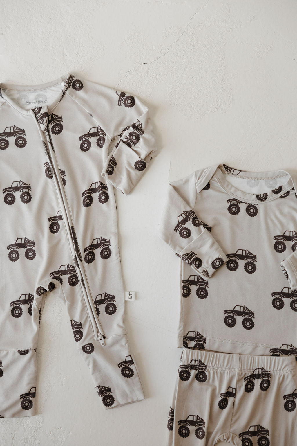 Two light gray Bamboo Zip Pajamas | Monster Truck by forever french baby are displayed flat. The left is a zip-up footed onesie; the right, a long-sleeve shirt with matching pants. Both feature black monster truck prints and breathable fabric.