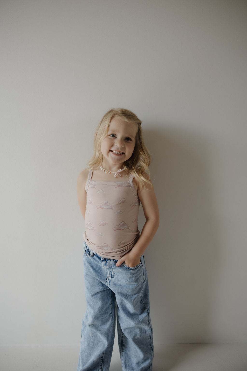 A young girl with long blonde hair smiles against a plain wall, wearing the Kendy 3 Toddler Bamboo Cami Set | Ruby—a light pink, sleeveless, breathable top with a cloud pattern—paired with loose blue jeans and hands in her pockets.