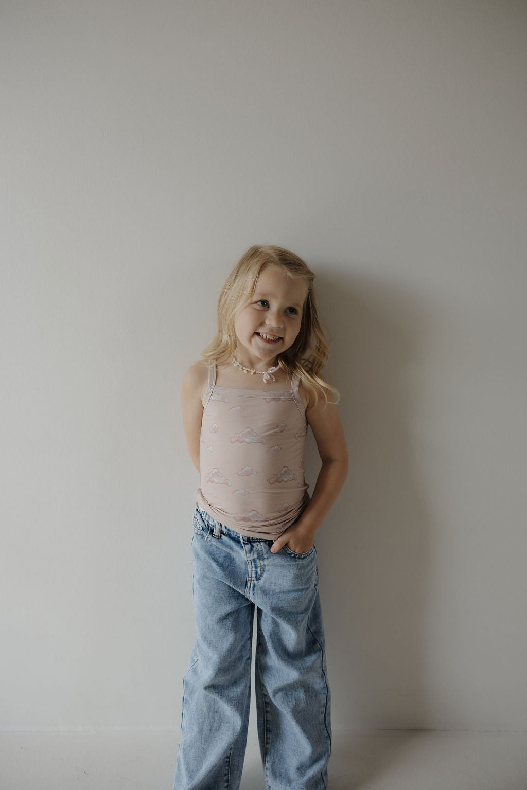 A young child with long blonde hair, smiling with one hand in their pocket, wears the Kendy 3 Toddler Bamboo Cami Set in Ruby—featuring a breathable pink tank top and loose blue jeans—standing against a light-colored wall.