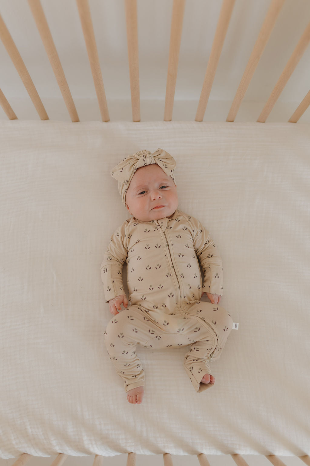 A baby contently lies in a crib on a white sheet, wearing forever french baby's Bamboo Zip Pajamas in the Flora pattern, with matching headband sporting small leaf designs.