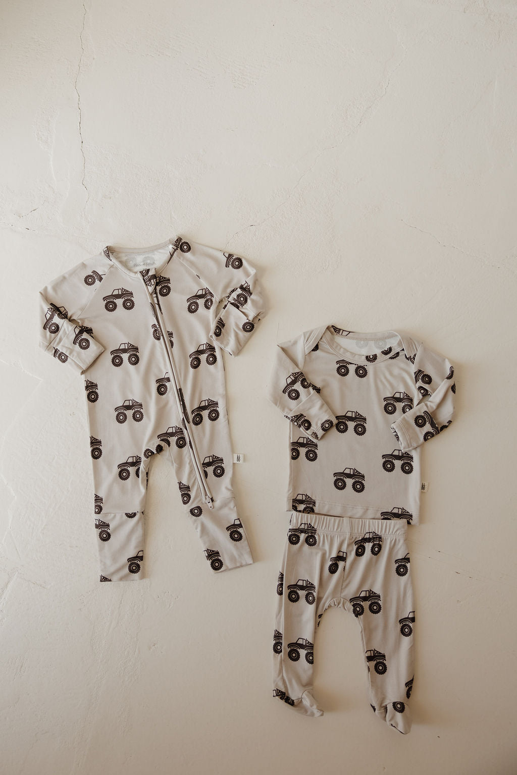 Two hypo-allergenic Sleepy Time Set | Monster Truck outfits from forever french baby, in beige bamboo with black car prints, are displayed: one zip-up onesie and one long-sleeve top with matching pants, ideal for sensitive skin.