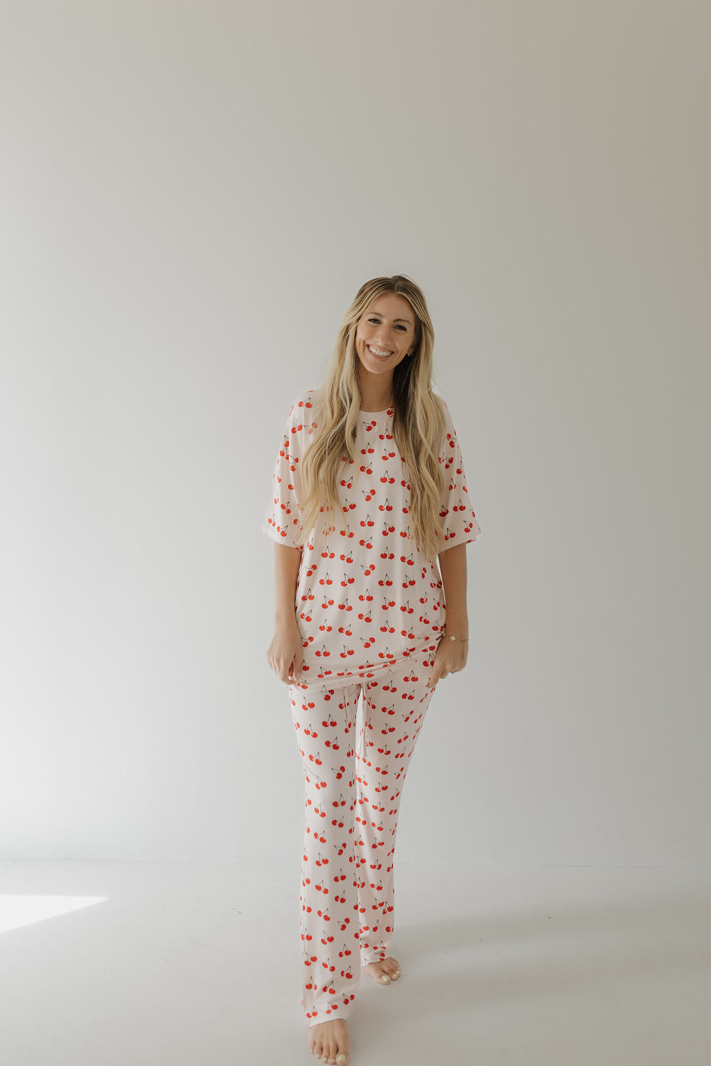 In a minimalist room with a plain background, a person smiles while barefoot, wearing forever french baby's Short Sleeve Women's Bamboo Pajamas in Cherry Orchard. Their long hair flows freely, creating a calm and relaxed atmosphere reminiscent of hypo-allergenic clothing ads.