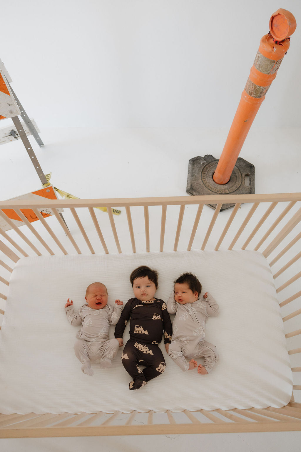 Three babies rest in a wooden crib on a white sheet; two wear light gray forever french baby Sleepy Time Set | Excavator bamboo outfits while one wears a dark, patterned set. Outside the crib, an orange traffic cone and ladder appear in the hypo-allergenic room.