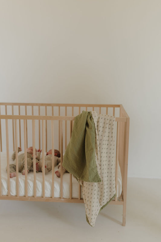 A baby rests in a light wooden crib with a white mattress. The forever french baby Muslin Quilt | Flora (Mis-Print Peach Tone) and a cream reversible muslin blanket with petite print hang over the rail against a pale wall.