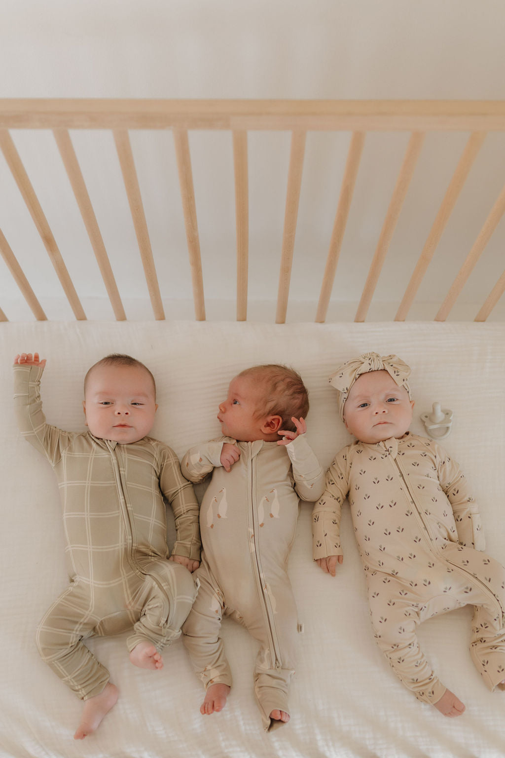 Three babies in matching neutral-toned onesies lie side by side in a crib. The left baby wears a plaid outfit, the middle one is in plain attire, and the right baby has a patterned onesie with a headband. The crib features wooden slats. The middle baby's outfit is from forever french baby's Bamboo Zip Pajamas | Linen Grid collection.