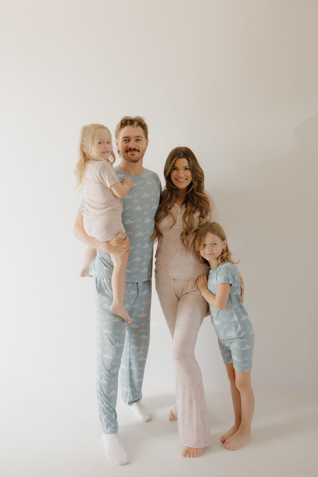 A family of four smiles in matching pastel pajamas against a light background. The mom wears Kendy 3’s Women's Bamboo Pajama | Ruby—ideal for breastfeeding moms and gentle on sensitive skin thanks to its hypo-allergenic fabric.