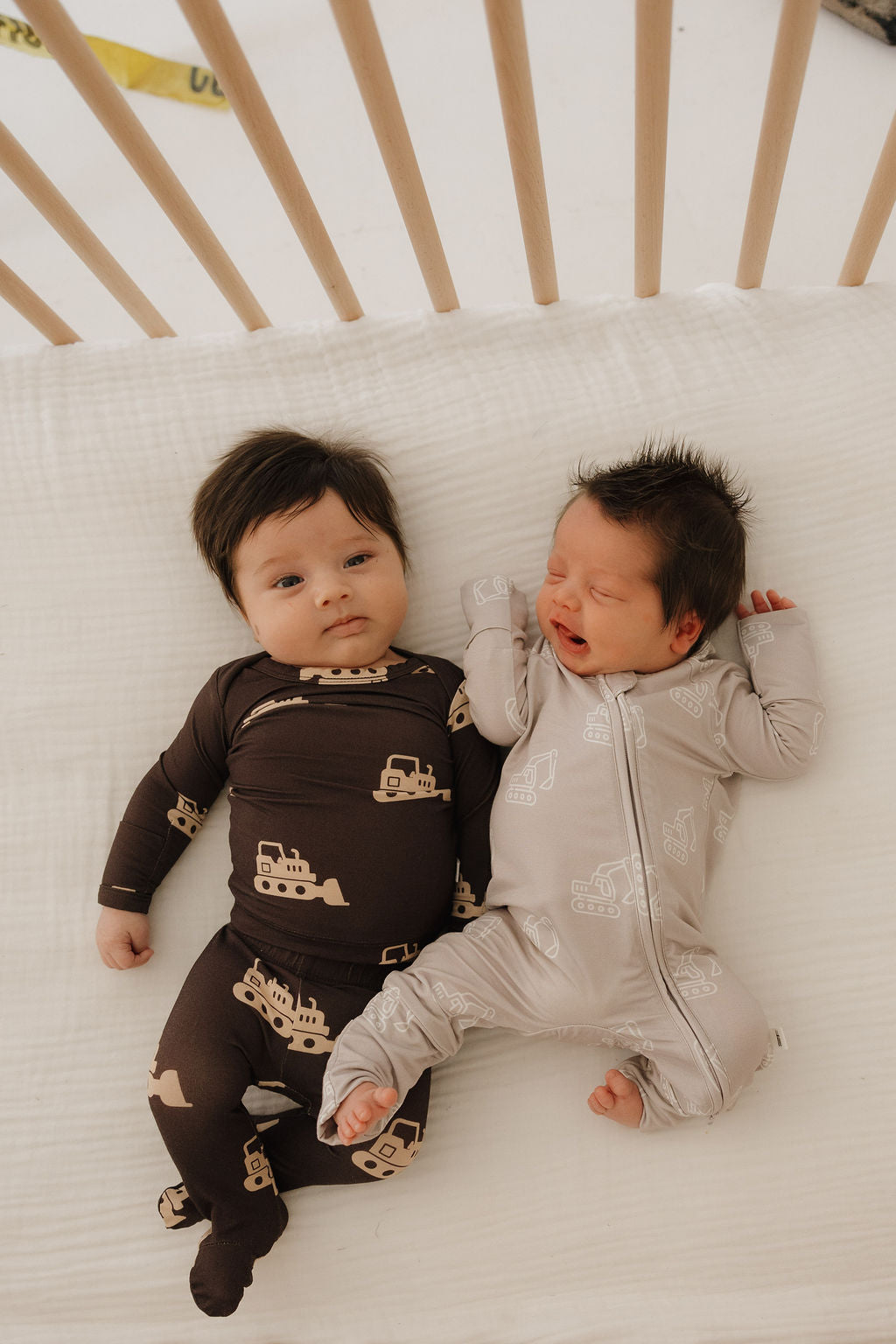 Two babies lie side by side in a crib: one wears forever french baby's Bamboo Zip Pajamas | Excavator with vehicle prints, looking at the camera; the other in light gray pajamas, both cozy in soft, breathable bamboo fabric.