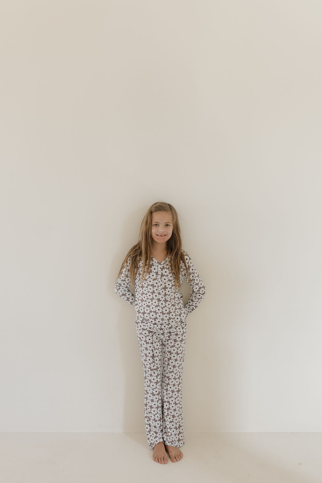 A young girl with long hair, smiling with her hands behind her back, stands barefoot against a plain white background, wearing matching floral-patterned Youth Flare Bamboo Pajamas by forever french baby.