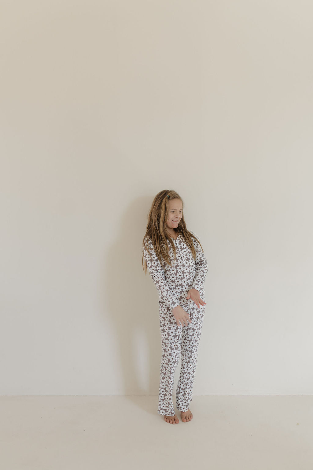 A young girl with long hair stands barefoot against an off-white wall, wearing forever french baby's Youth Flare Bamboo Pajamas in a floral pattern. Crafted from breathable bamboo fabric, she smiles with her hands together and looks to her right.