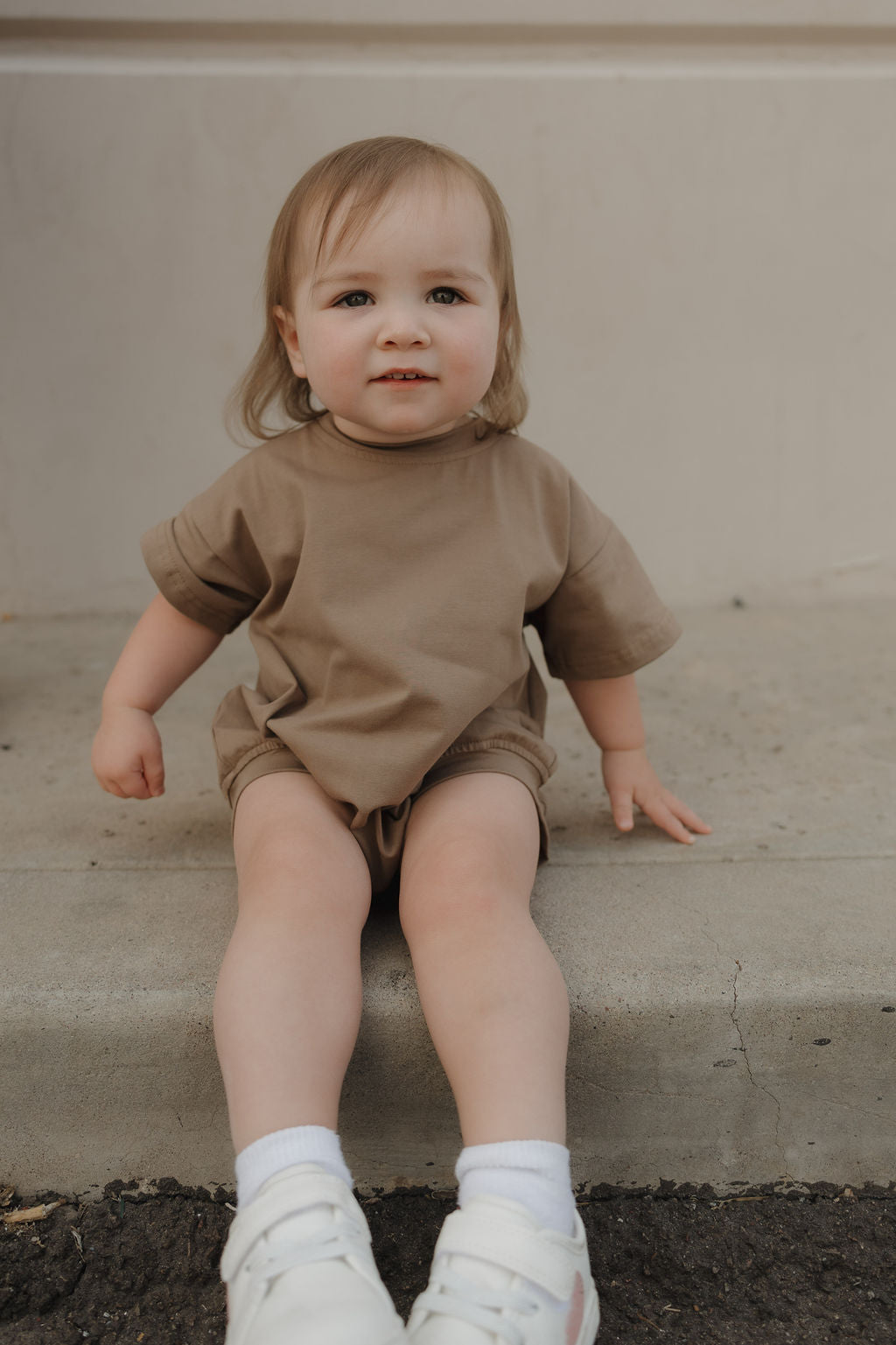 A toddler with light hair sits on a concrete step, smiling slightly at the camera while wearing the forever french baby Short Sleeve Romper in Patina and white sneakers.