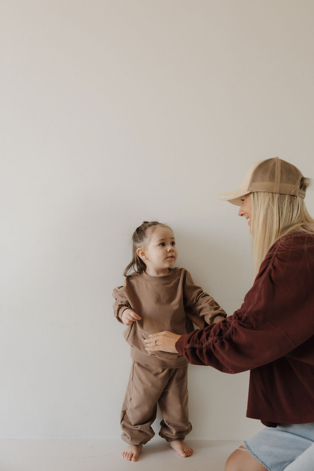 A barefoot young child wearing the forever french baby Child Sweat Set | Hot Cocoa stands against a plain light wall, looking at an adult woman in a cap and maroon sweatshirt who kneels nearby, smiling and reaching out.