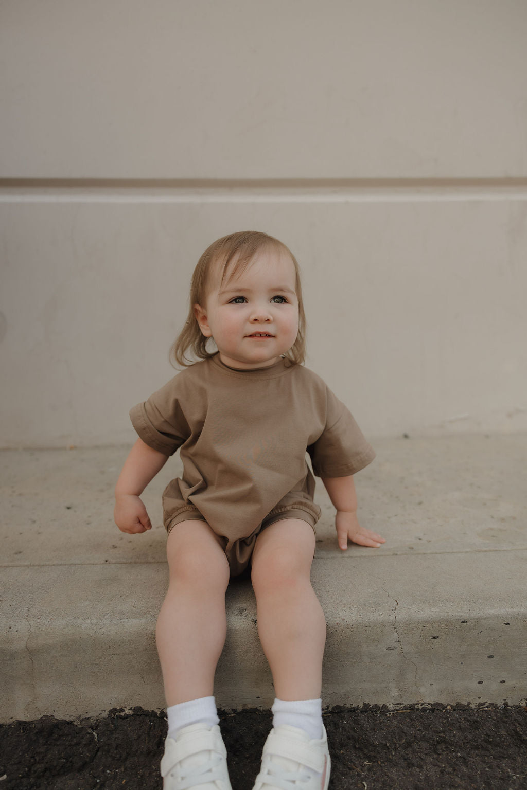 A toddler with light hair sits on a concrete step wearing the forever french baby Short Sleeve Romper in Patina, featuring a relaxed fit and snap closure, paired with white shoes and socks, against a plain wall background.