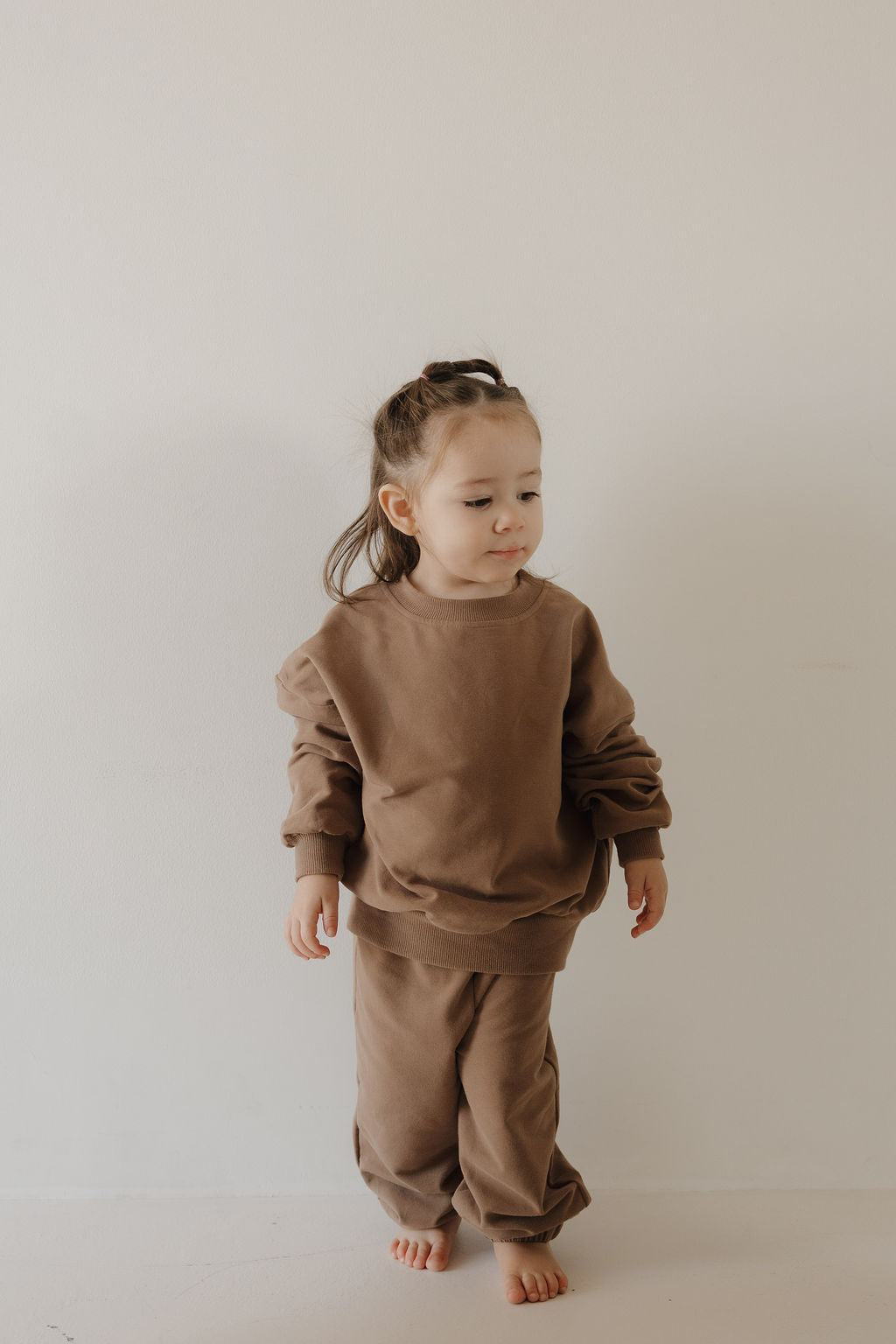 A young child with light brown hair stands barefoot against a plain off-white background, wearing the forever french baby Child Sweat Set in Hot Cocoa—a loose brown sweatshirt and matching sweatpants.