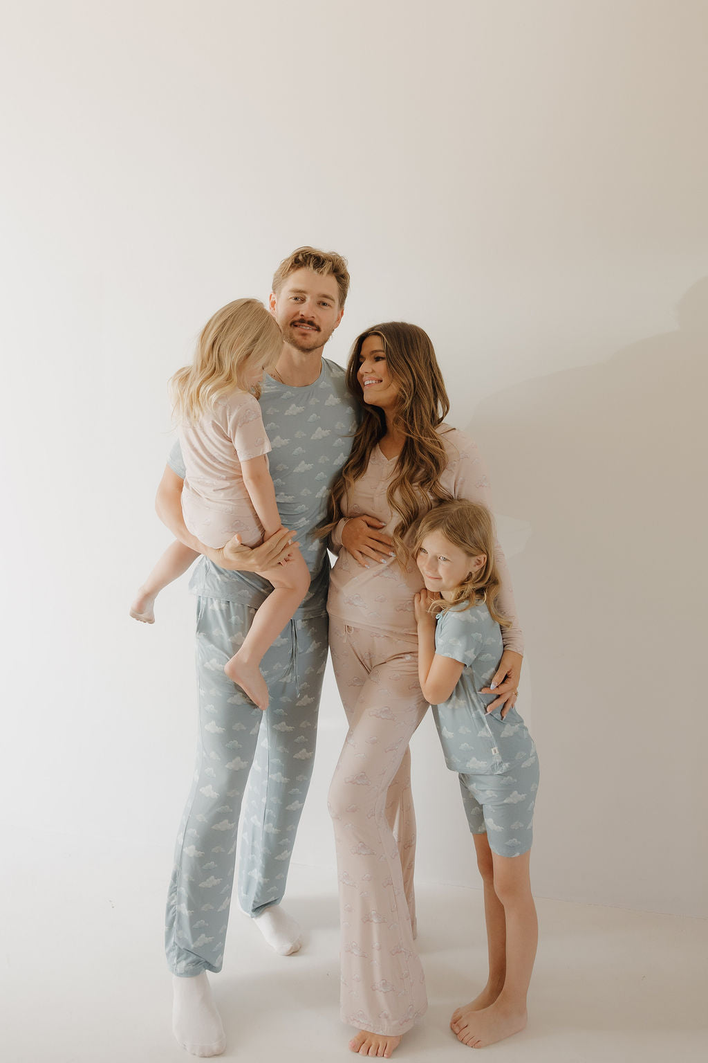 A smiling family of four poses barefoot in breathable Kendy 3 Bamboo Short Set | Ruby pajamas. Dad holds a child, while the pregnant mom stands beside him with another child; all wear matching pastel sleepwear against a light background.